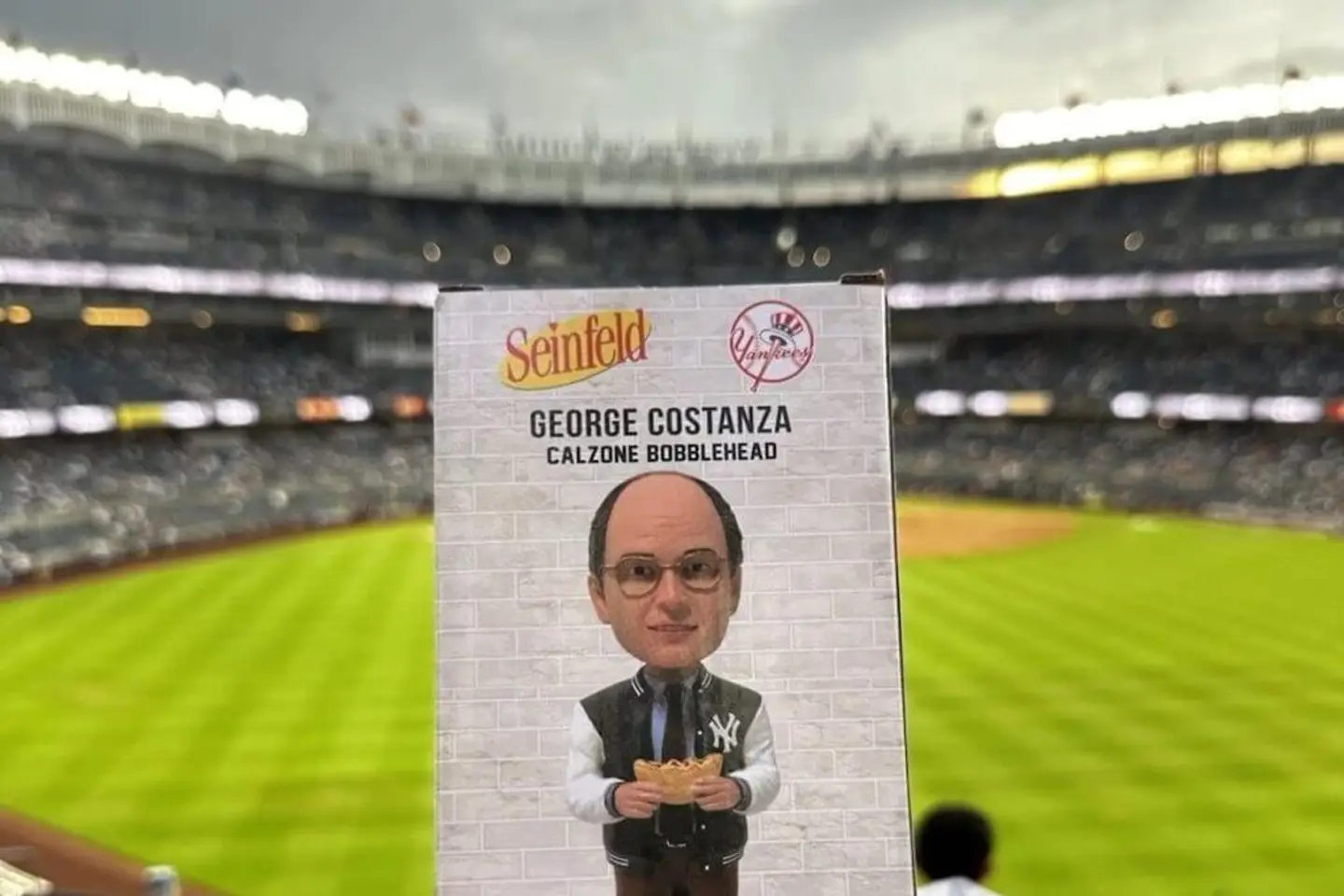 George Costanza bobblehead in a Yankees letterman jacket holding a half-eaten calzone